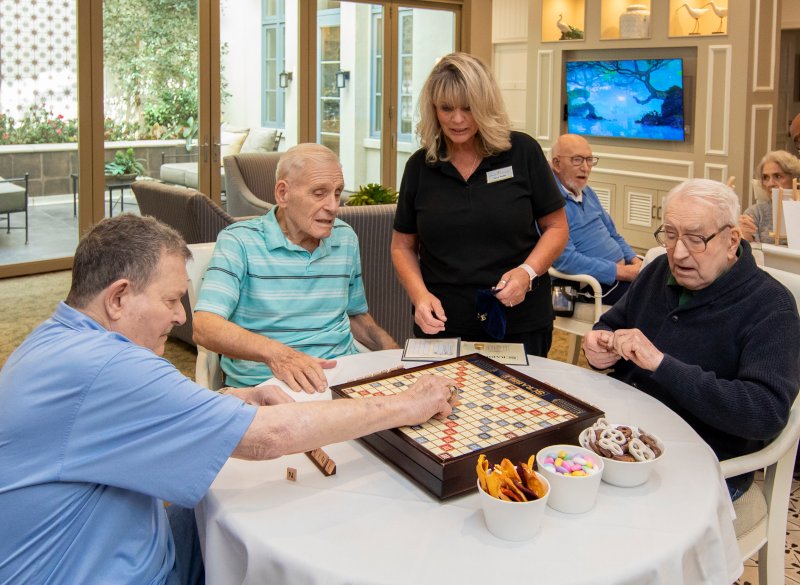 3 elderly men playing Scrabble while a care assistant gives advice.